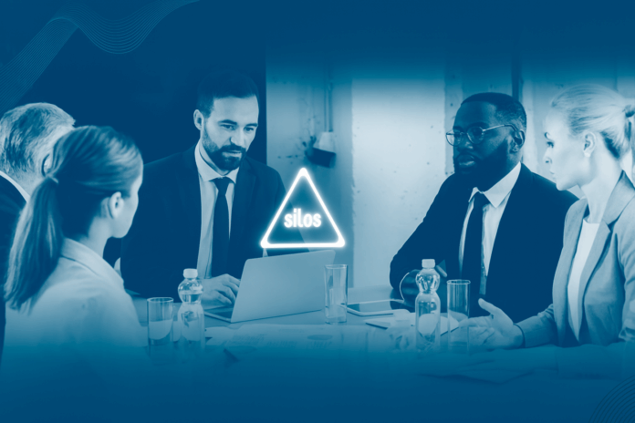 Executivos em reunião com alerta de silos na tela do laptop.
