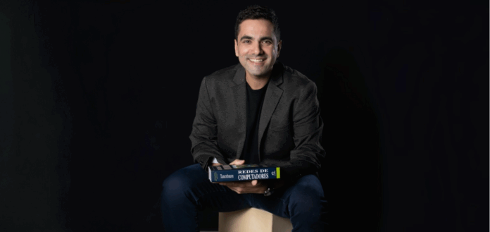 Autorregulação em inteligência artificial representada por profissional de tecnologia sorridente, sentado em banco de madeira, segurando o livro “Redes de Computadores” sobre o colo, com fundo preto.
