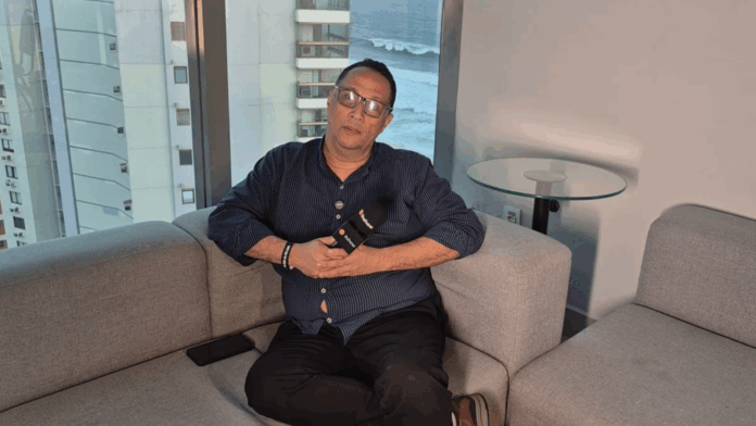 SN Informática representada por executivo que está sentado em um sofá durante uma entrevista, segurando um microfone com a logomarca do portal Itshow. Ao fundo, há uma janela ampla com vista para prédios e o mar.
